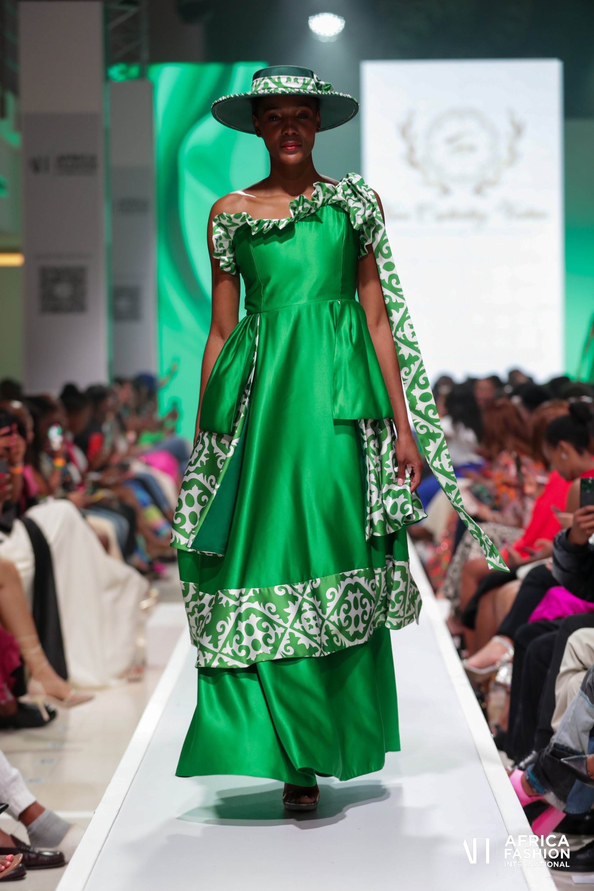 A model wearing the Tumi Captivating Green Dress. The dress features a captivating green and white print on luxurious printed satin. The model is shown posing gracefully, highlighting the flowy tiered-flare silhouette.